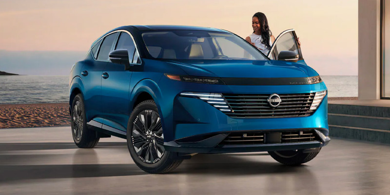 2026 Nissan Murano SUV in blue parked near a beachfront setting at sunset, featuring a sleek modern design, wide grille, and a woman stepping out of the driver’s side door.